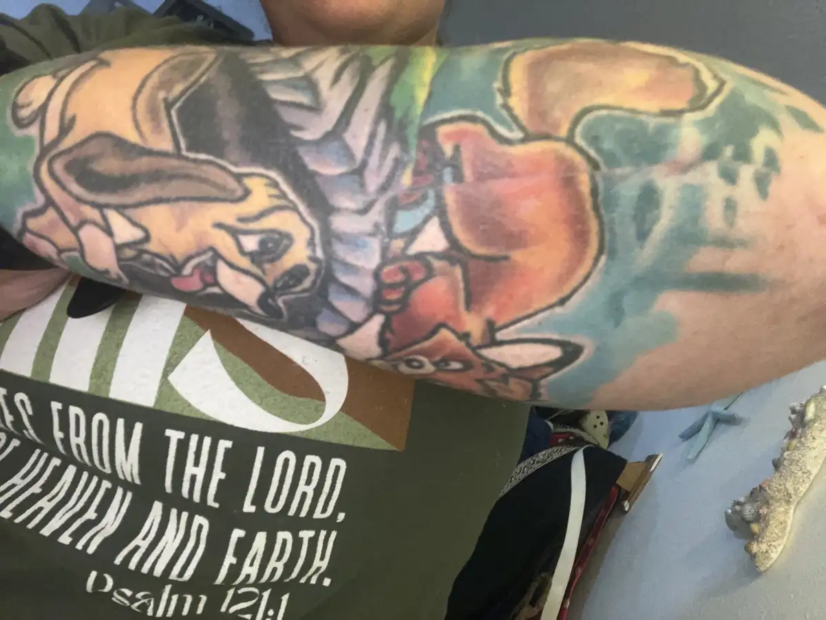 A person's tattooed forearm displays a colorful, cartoon-style tattoo of two dogs, one tan and one reddish-brown, facing…