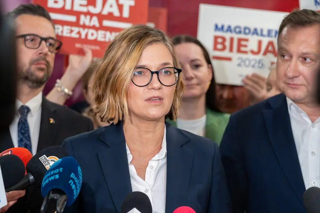 A woman with short blonde hair and black-rimmed glasses, wearing a dark blazer over a white shirt, stands at a press…