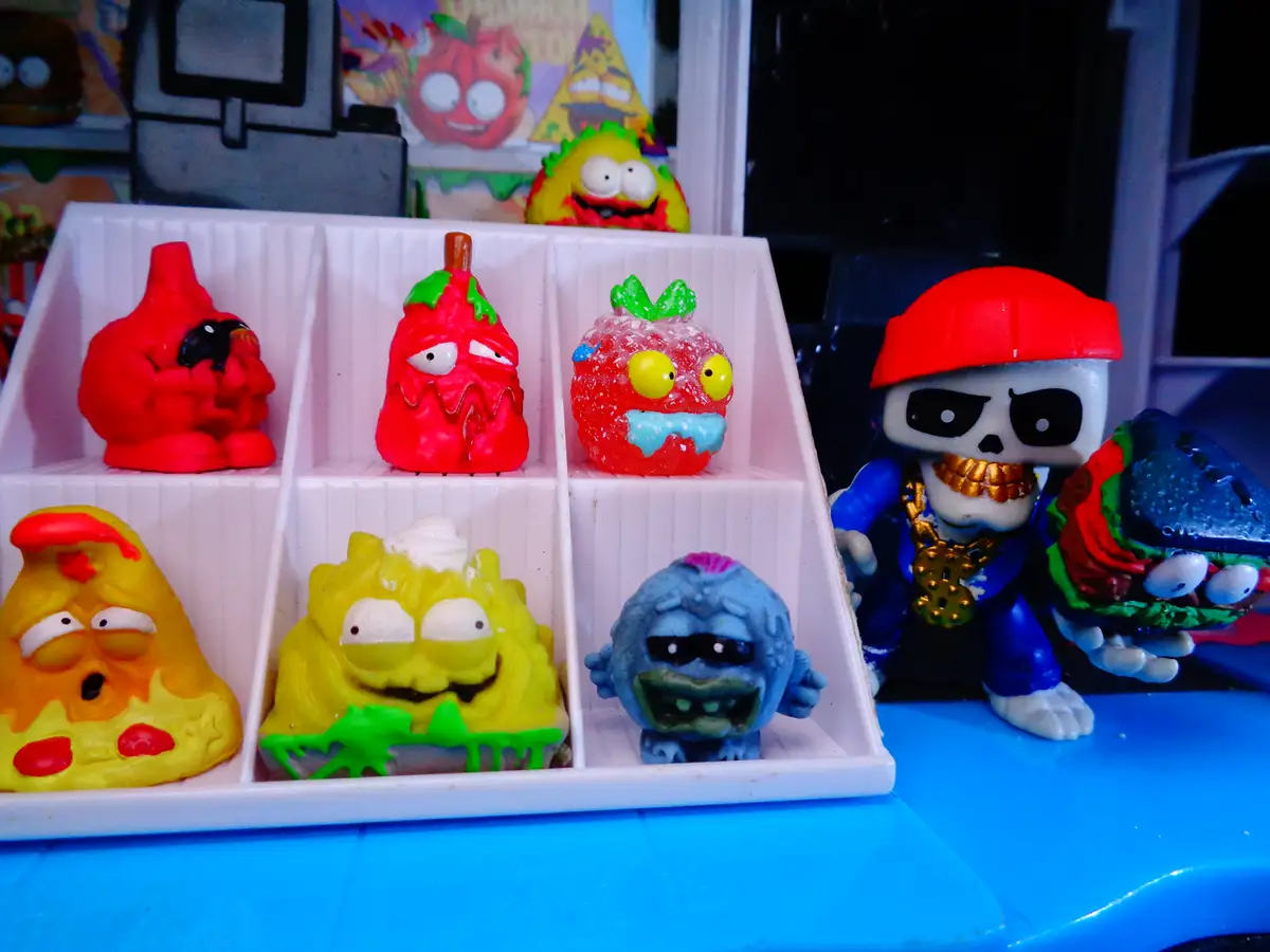 A white plastic tray holds six small, colorful, grotesque-looking toy figures shaped like food items such as a red tomato, a…