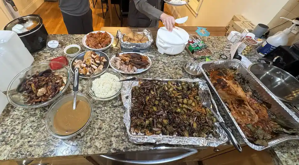A kitchen counter is covered with a large spread of holiday-style food including roasted turkey, mashed potatoes, stuffing…