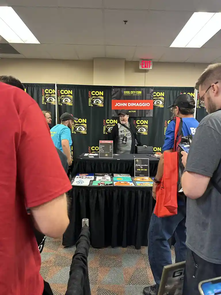 A man identified as John DiMaggio, an actor, stands behind a signing table at a convention booth labeled 'Steel City Con'…