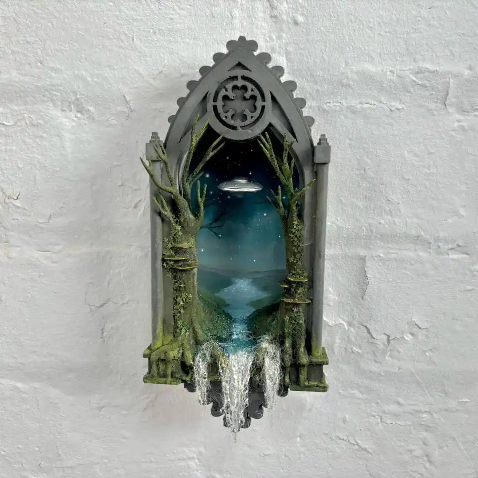 A 3D diorama mounted on a white wall depicts a gothic-style window frame with moss-covered pillars and bare trees. Through…