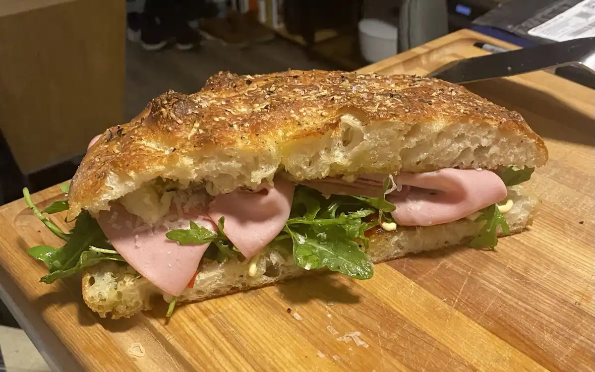 A close-up photo of a freshly made sandwich on crusty artisan bread, filled with pink deli meat, green arugula, and shredded…