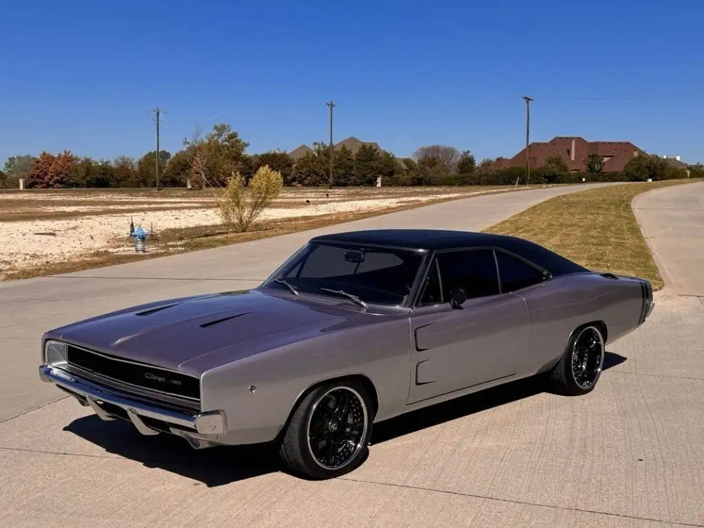 a silver dodge charger with a black roof and black wheels is parked on a concrete driveway under a clear blue sky. houses…