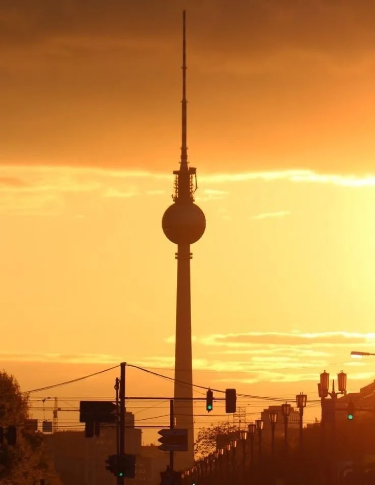A silhouette of the Berlin TV Tower against a vibrant orange sunset sky, with streetlights and traffic signals visible in…