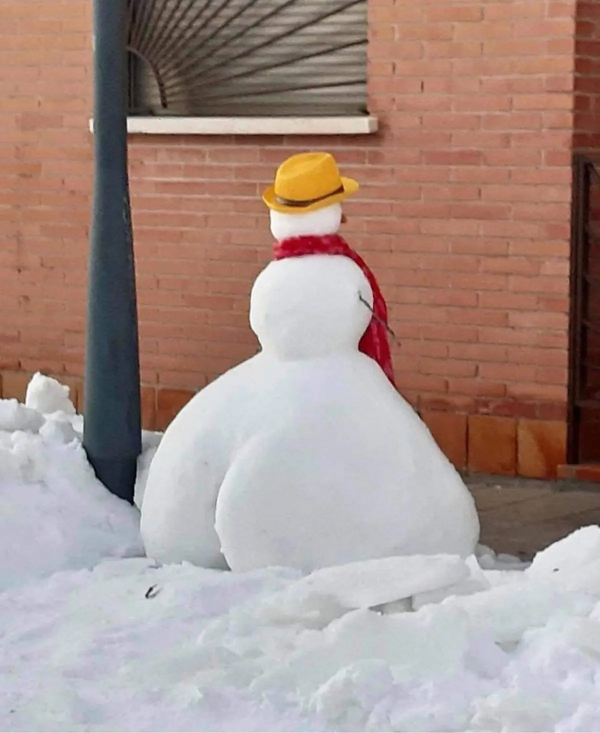 A snowman viewed from behind, wearing a yellow hat and red scarf, sits in a snowy area next to a brick building and a dark…