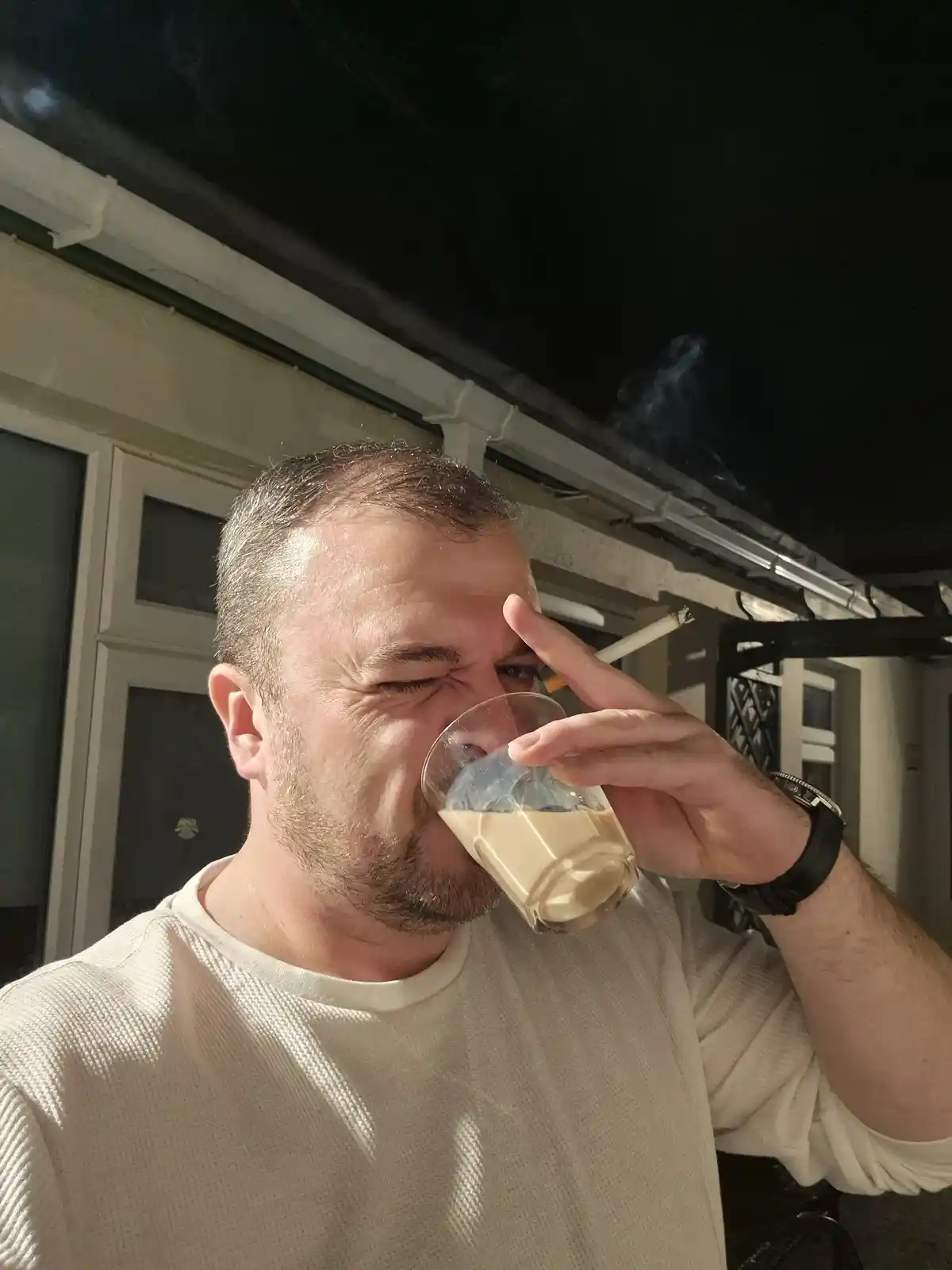 A man with short dark hair and a beard, wearing a white t-shirt, is drinking from a glass of light-colored liquid while…