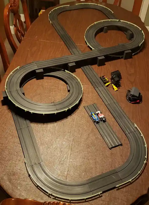 A plastic slot car racing track is laid out in a complex, looping pattern on a wooden dining table. Two small slot cars sit…