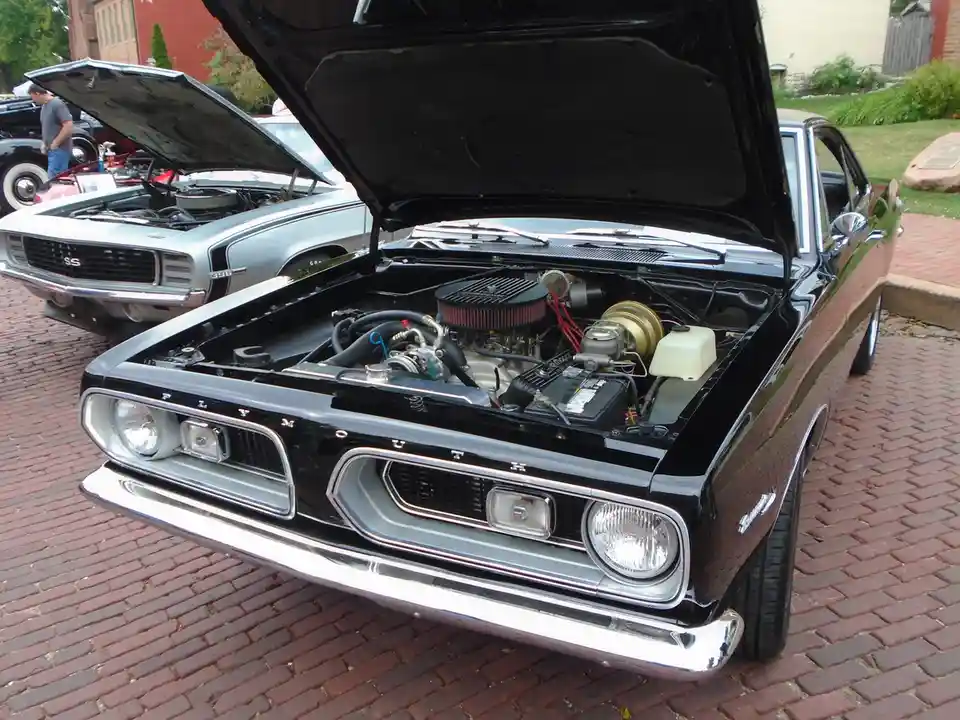 A black classic Plymouth car with its hood open, revealing a detailed engine bay, parked on a brick surface. In the…
