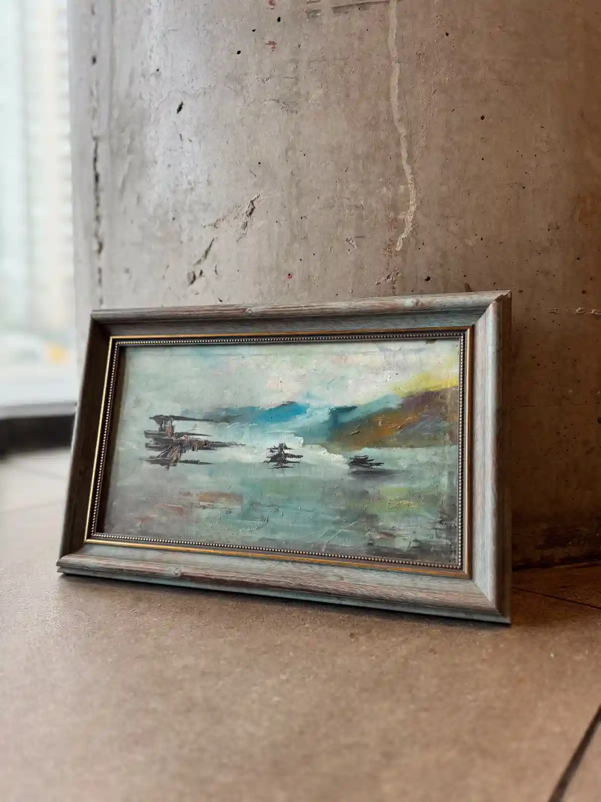 A small, framed painting of three stylized, abstracted boats on water rests on a tiled floor against a concrete wall. The…