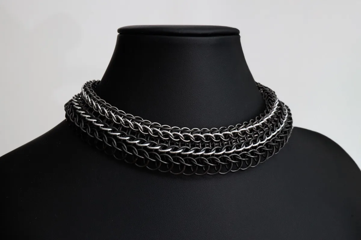 A black mannequin bust displays a thick, multi-layered chainmail choker necklace against a plain white background. The…