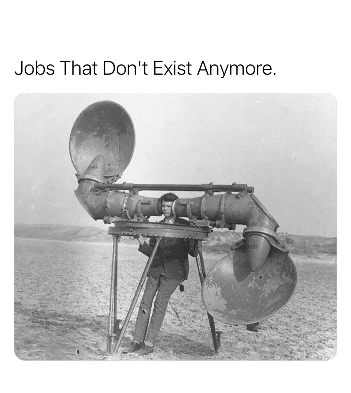 A black-and-white historical photo shows a man standing behind a large, complex, and outdated acoustic device with two…