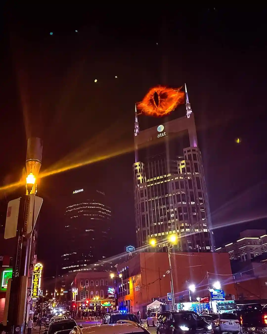 A nighttime cityscape photograph of a downtown area with illuminated buildings and streetlights, featuring a large, fiery…