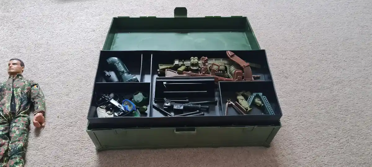 A toy soldier in camouflage uniform lies on a carpet next to an open green military-style toolbox filled with various toy…