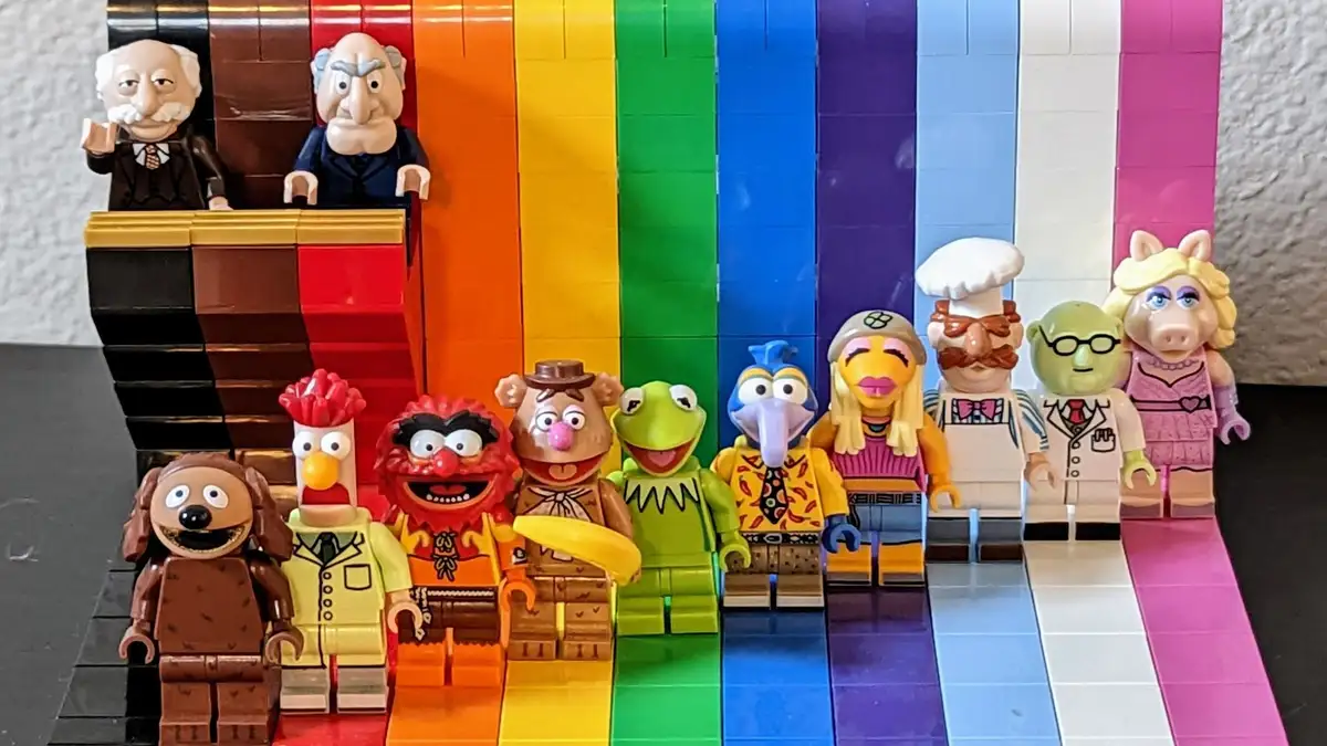 A lineup of LEGO-style Muppet minifigures stands on a rainbow-colored LEGO platform, with two older male figures seated at a…