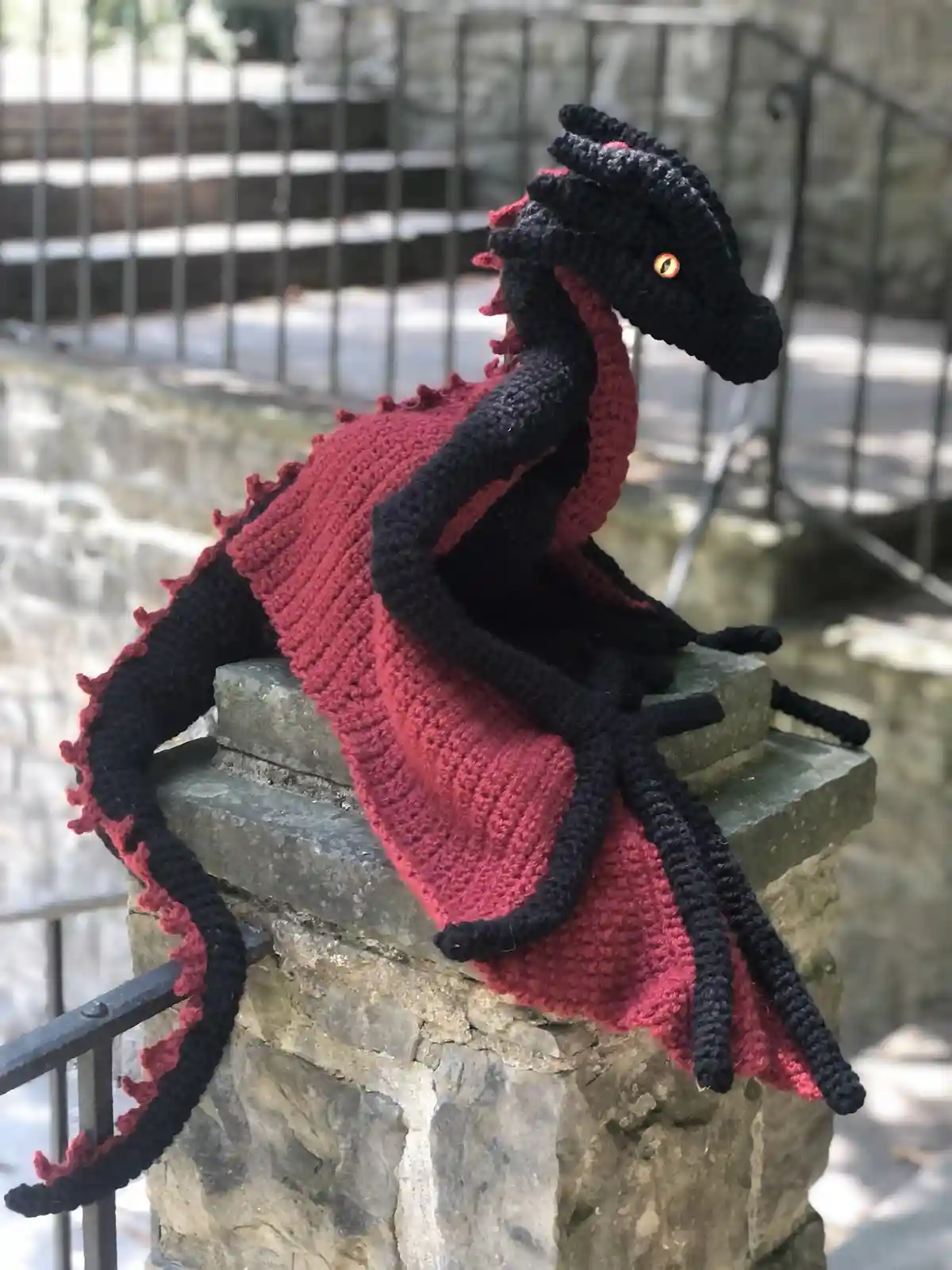 A hand-knitted dragon plushie, primarily black with red accents on its wings and spines, sits perched on a stone pillar…