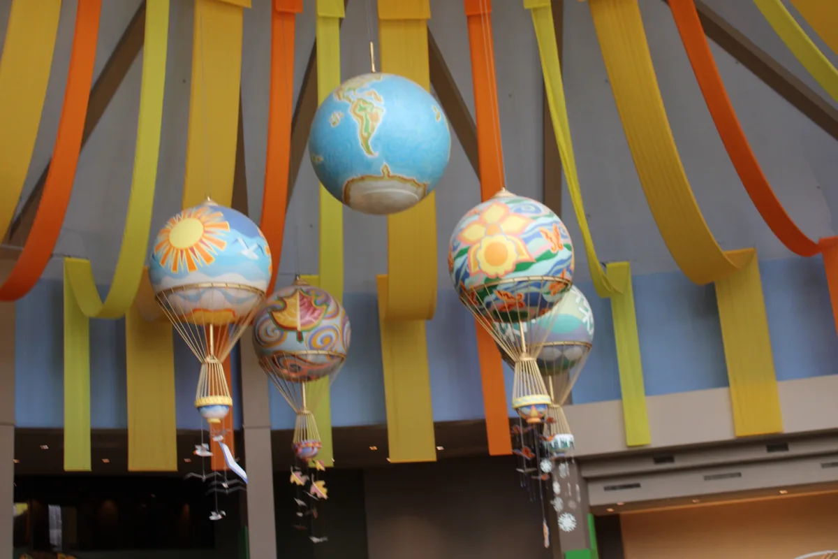Several colorful, decorative hot air balloon sculptures hang from a ceiling adorned with yellow and orange ribbons. One…