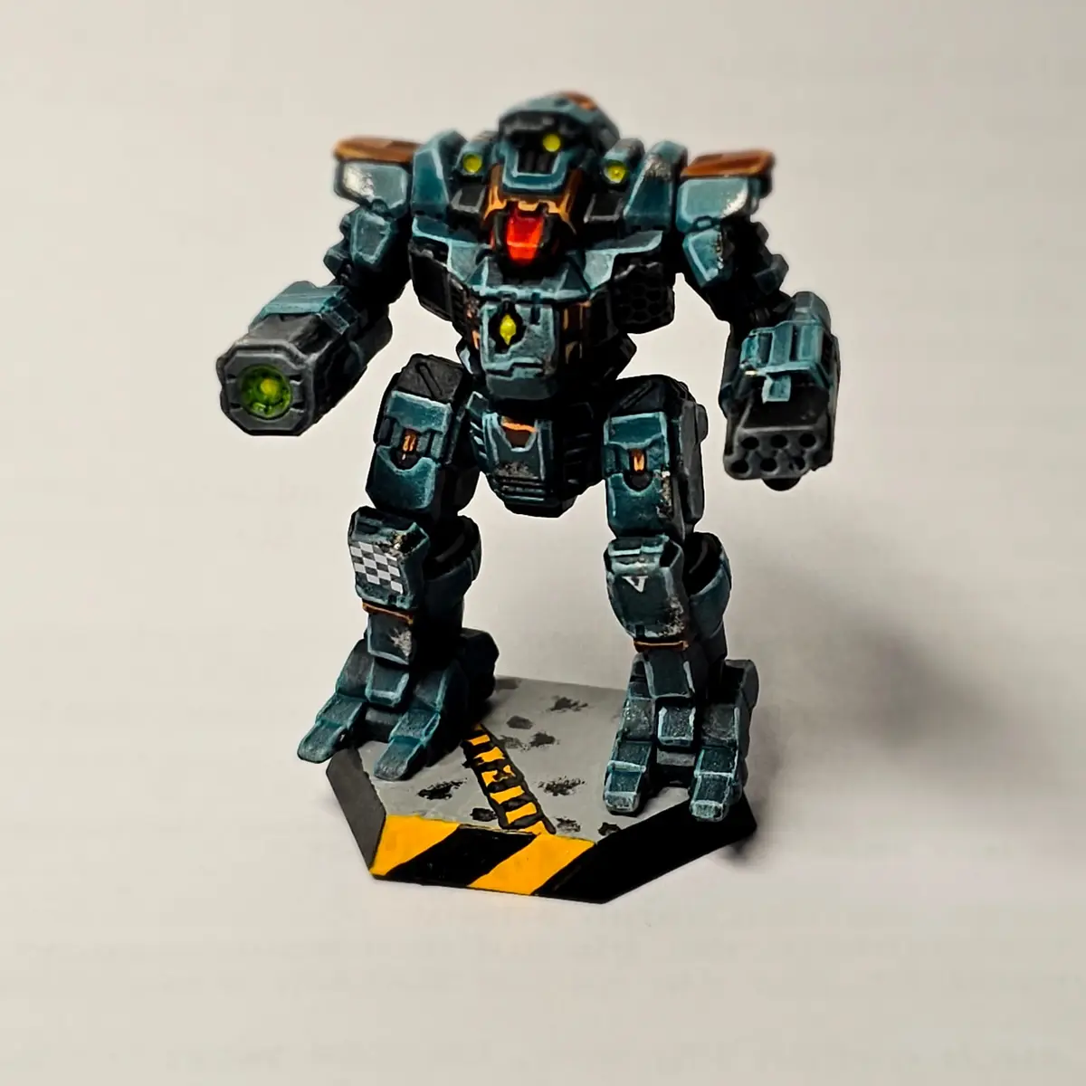 A painted miniature of a heavily armored, teal and black humanoid robot with glowing yellow eyes and red mouth, standing on…