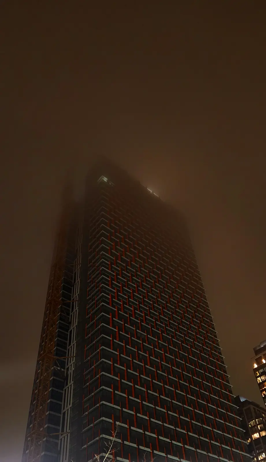 A low-angle nighttime photo shows a tall skyscraper partially obscured by thick fog, with its upper floors barely visible…