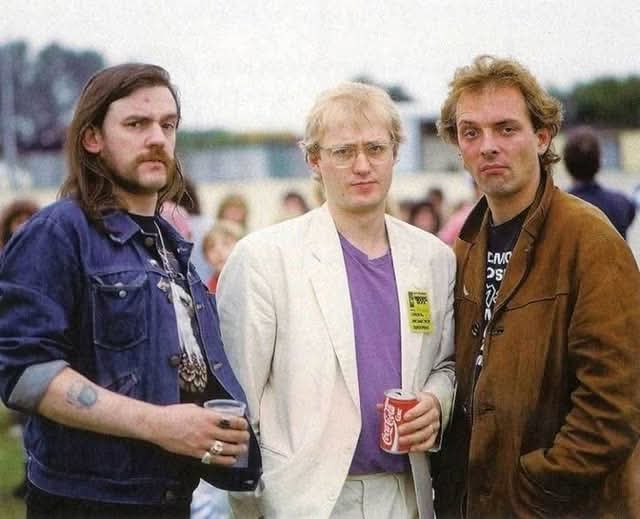 Three men stand side by side outdoors, each holding a drink. The man on the left has long hair and a mustache, wearing a…