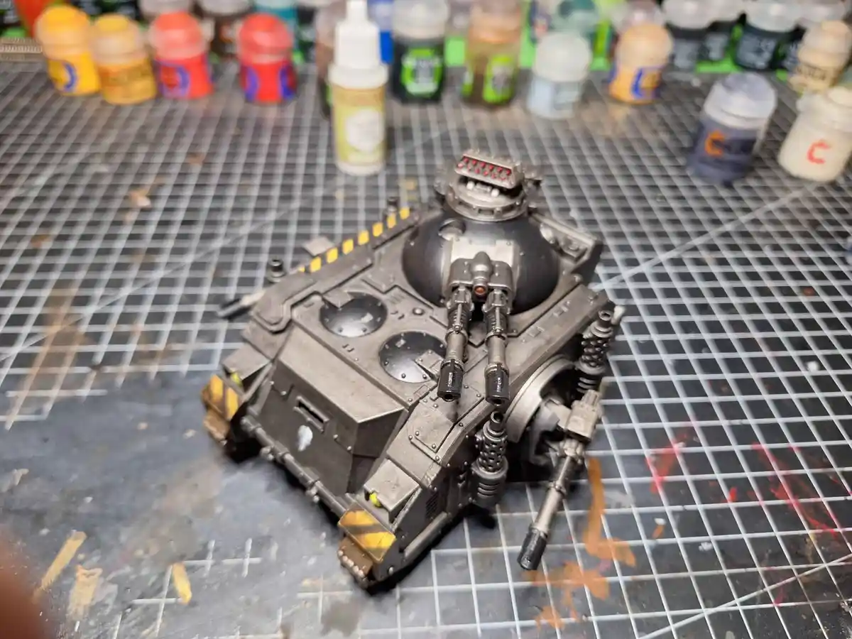 A detailed, unpainted miniature tank model sits on a gridded work mat surrounded by paint pots, with the overlaid text 'i…