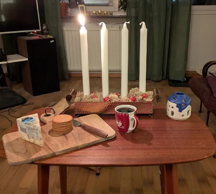 A cozy indoor scene shows a wooden coffee table with a lit advent candle set (one candle burning, three unlit), a red mug, a…