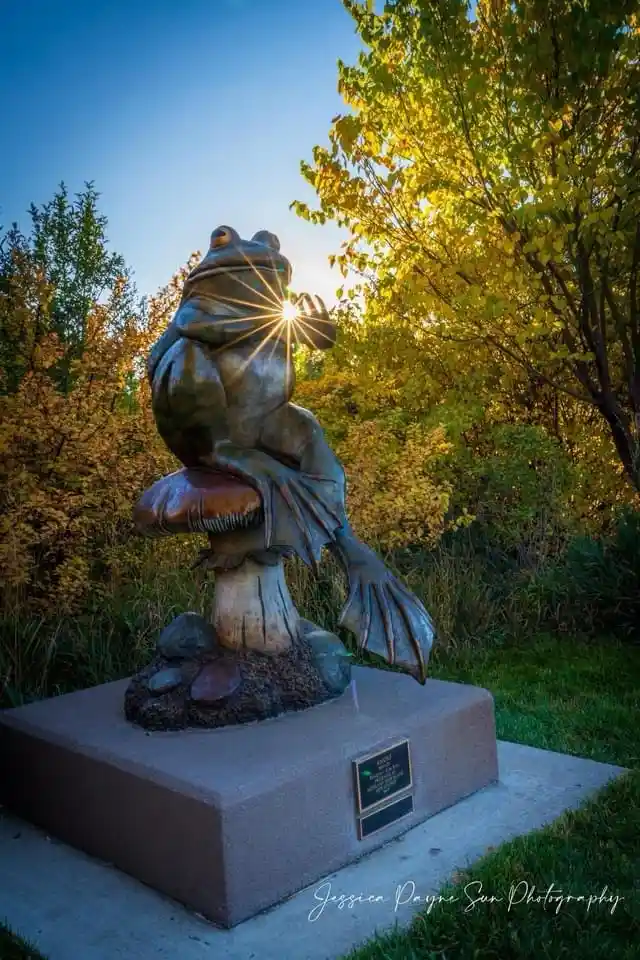 A bronze statue of a frog sitting atop a mushroom, with its front paws raised to frame the sun, creating a starburst effect…