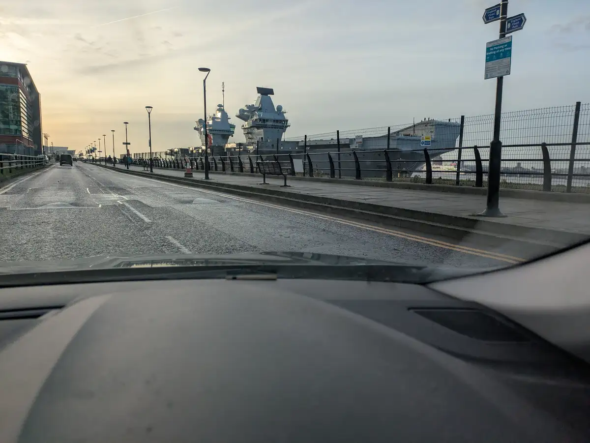 A photo taken from inside a car shows a road next to a harbor with large warships docked behind a fence. A signpost on the…