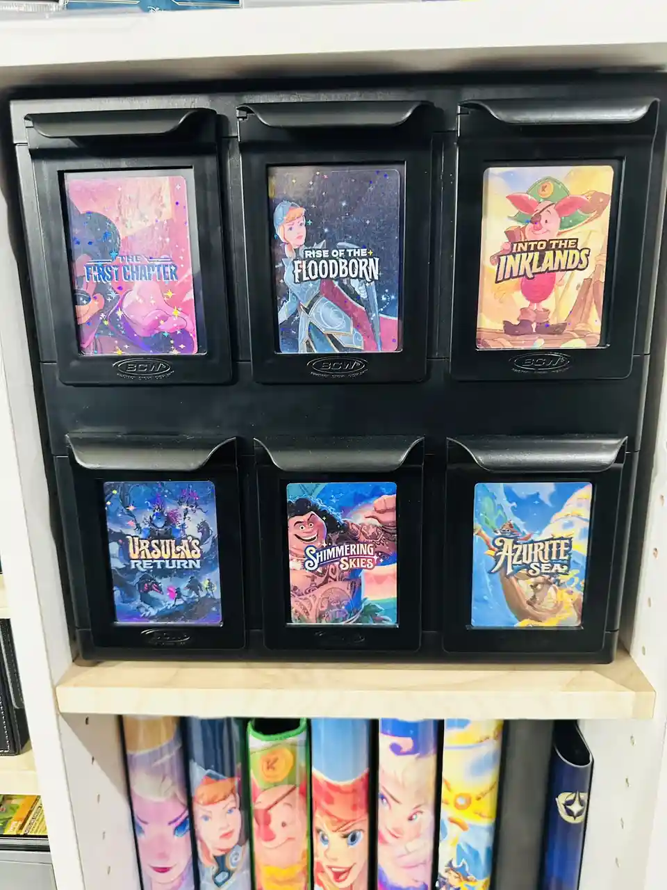 Six black BCW storage trays on a shelf display Disney-themed trading card game expansions with titles like 'The First…