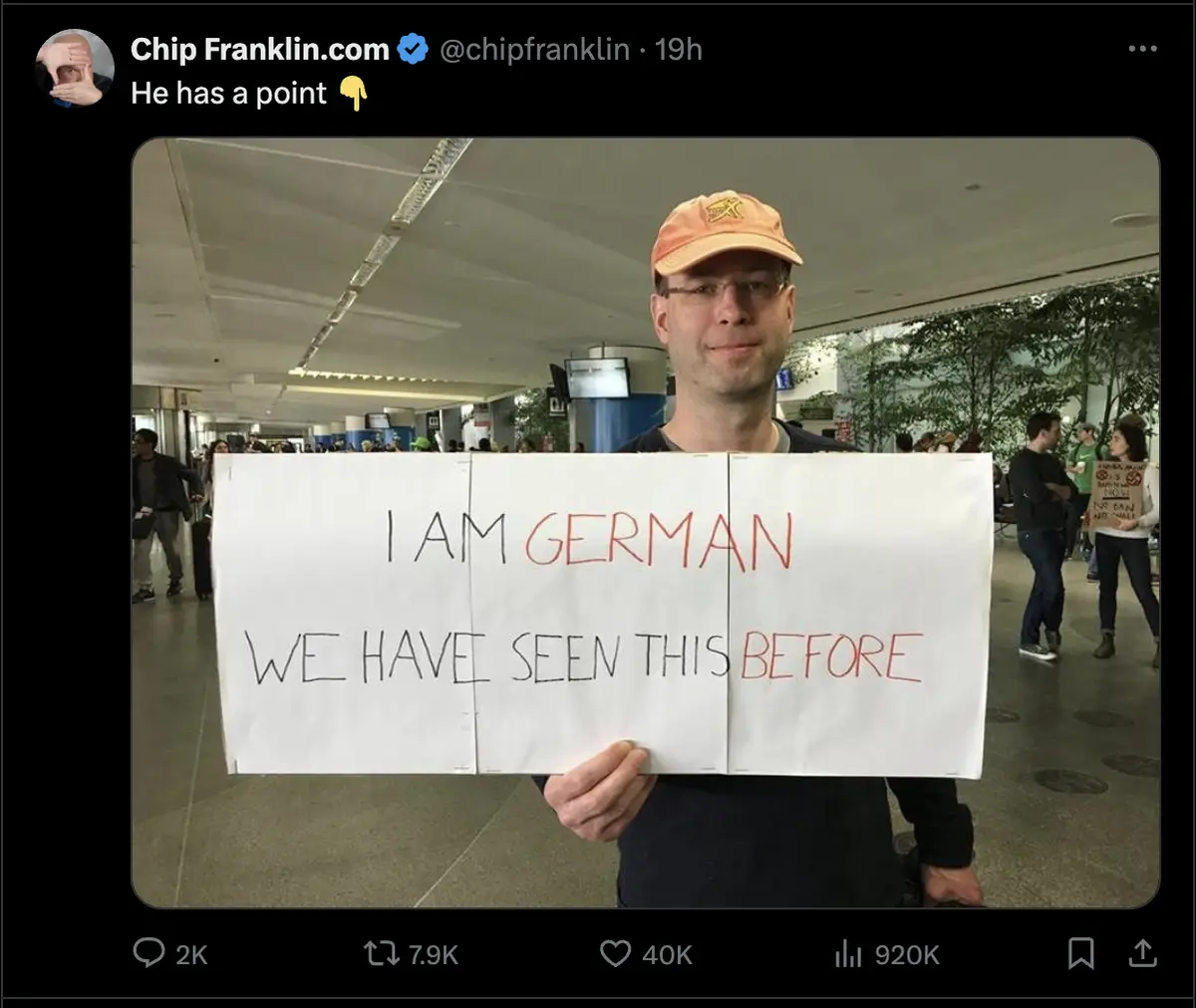 A man wearing glasses and an orange cap holds a large white sign in an airport terminal. The sign reads 'I AM GERMAN' in…