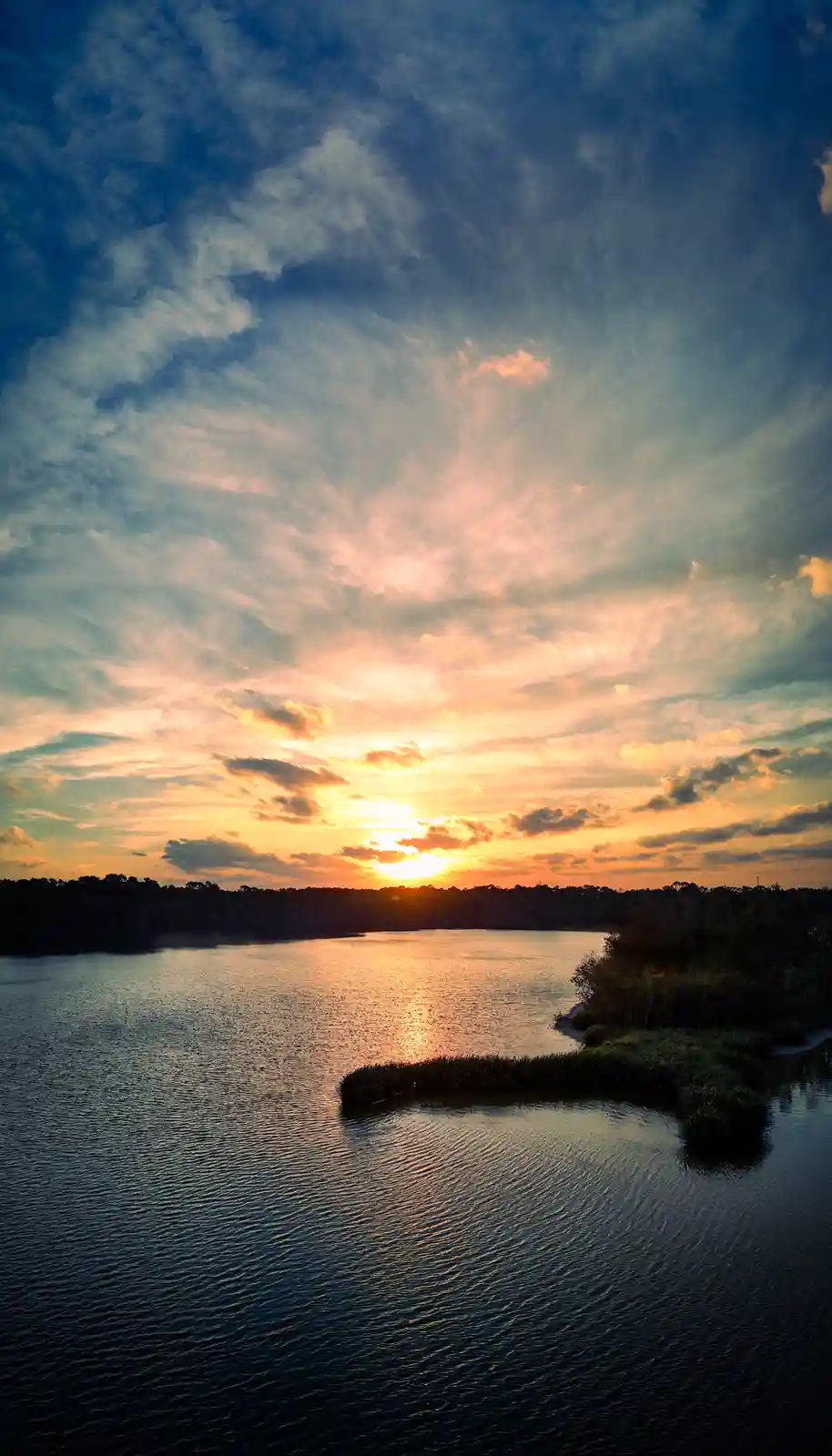 A serene sunset over a calm lake with ripples, surrounded by dark tree silhouettes under a sky painted with orange, pink…