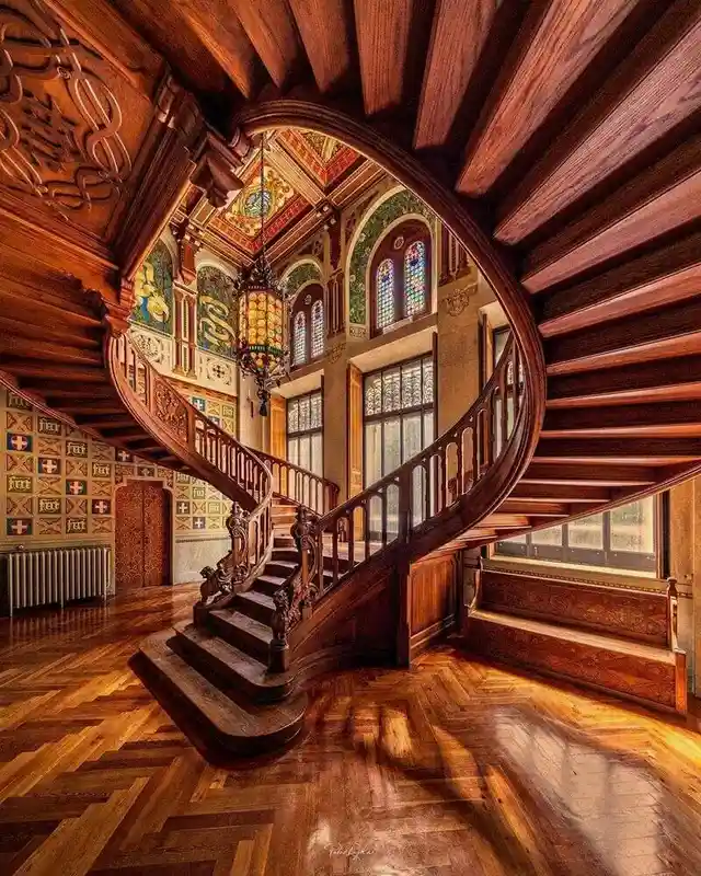 A grand, ornate wooden spiral staircase with intricate carvings and stained glass windows, bathed in warm sunlight on a…