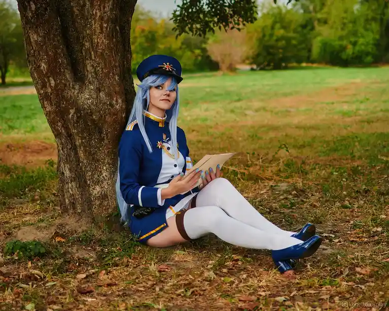 a person in a blue and white military-style cosplay outfit with light blue hair and a matching cap sits on the grass leaning…