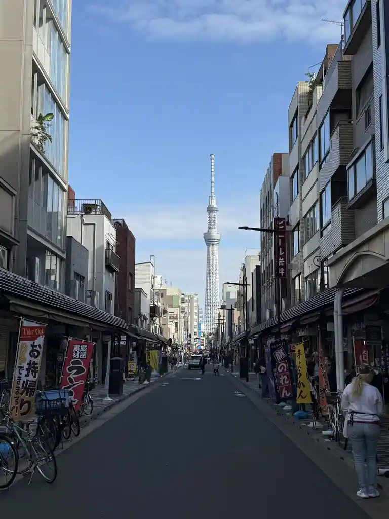 A narrow Tokyo street lined with buildings and shops, leading toward the distant Tokyo Skytree under a clear blue sky…