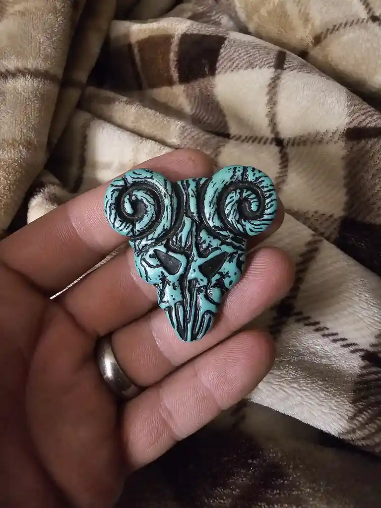 A hand holds a turquoise-colored, stylized ram skull pendant with black detailing, resting on a plaid blanket. The text…