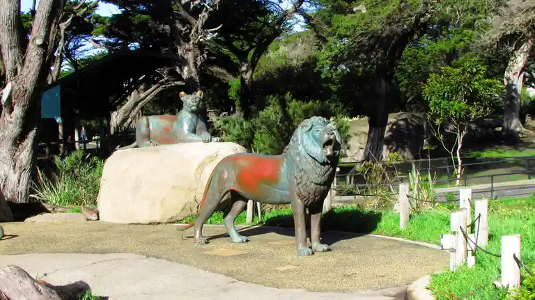 Two bronze lion statues in a park setting, one standing roaring and one lying down, both have red paint splotches on their…