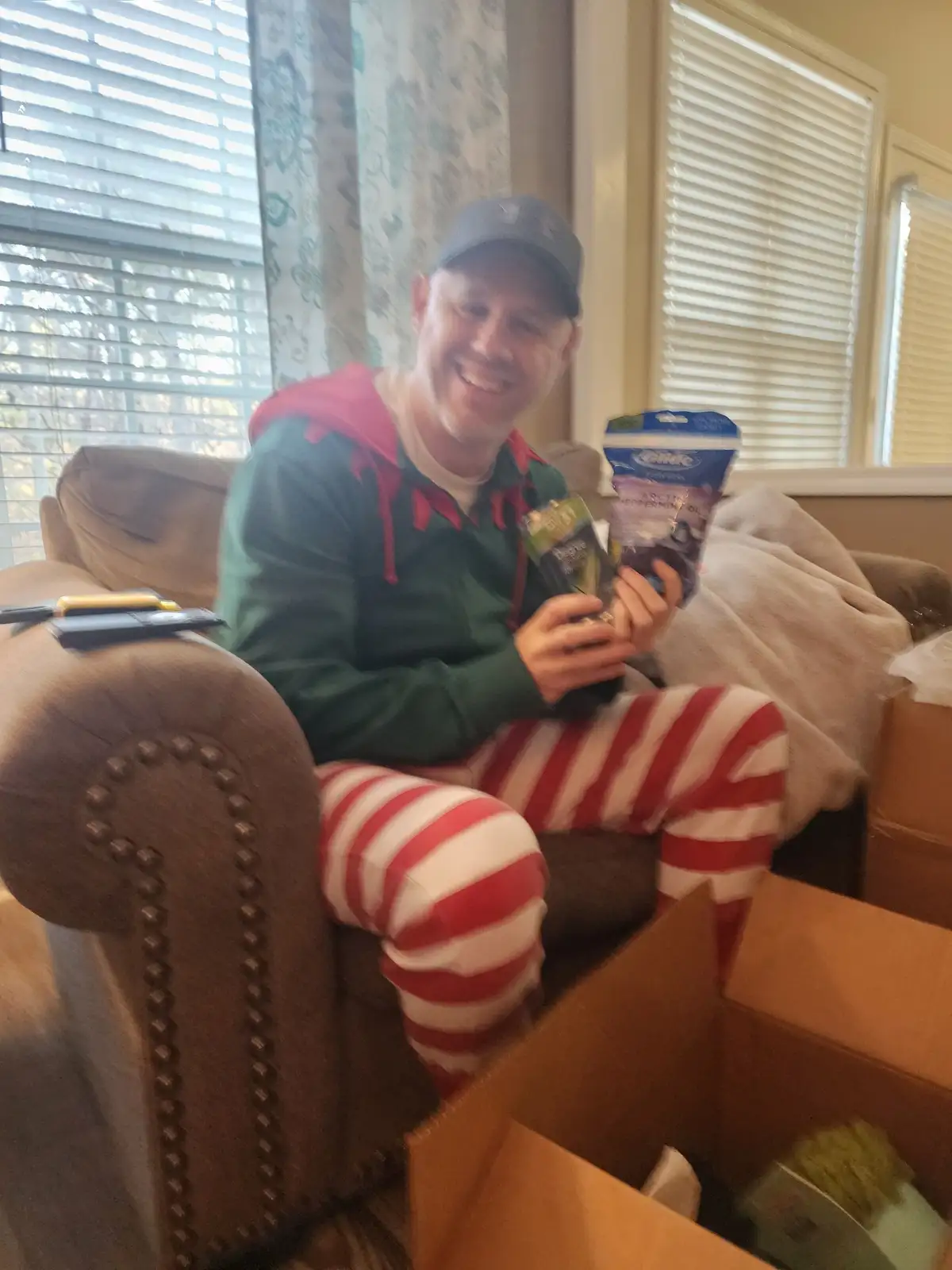 A man wearing green elf pajamas with red trim and red-and-white striped pants sits on a brown couch, smiling broadly while…