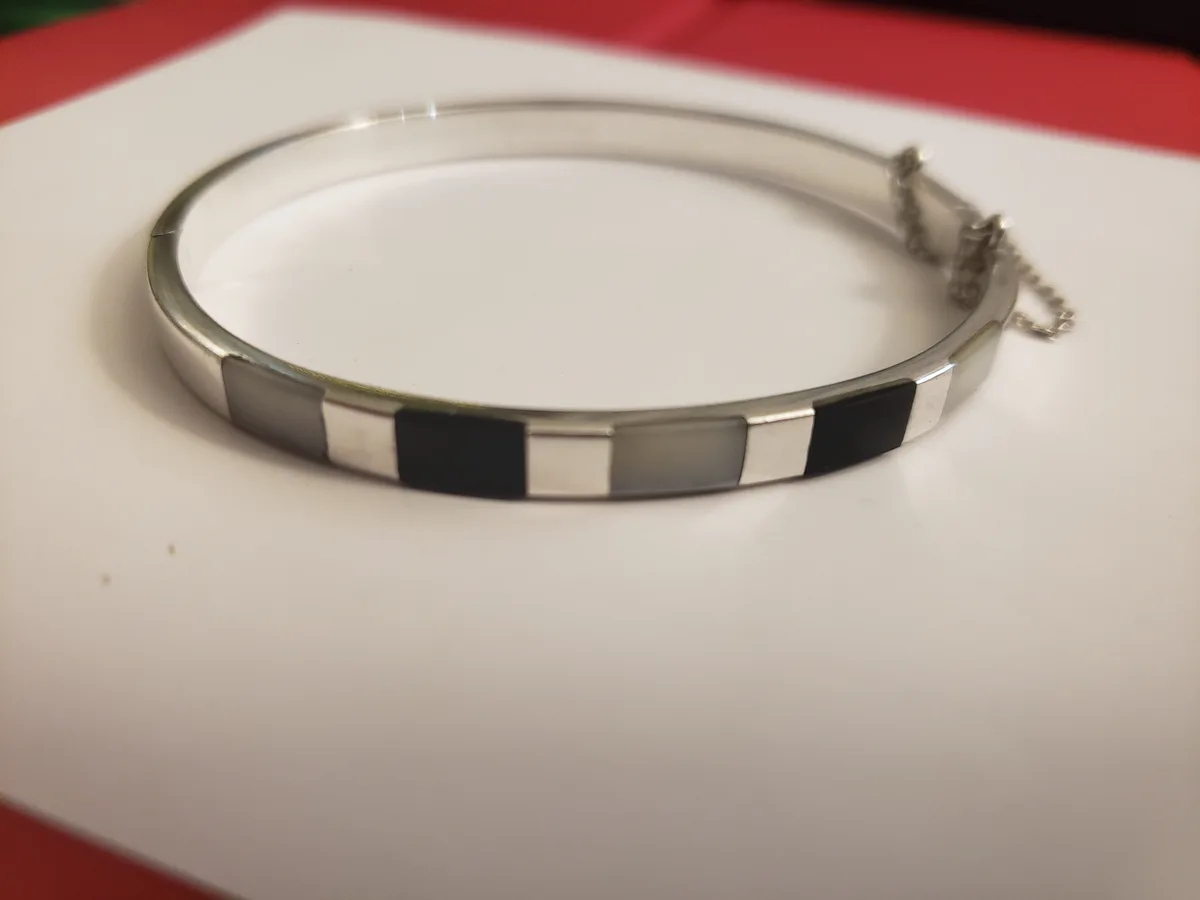 A silver bangle with alternating black and gray rectangular inlays rests on a white surface, with a red background. The…