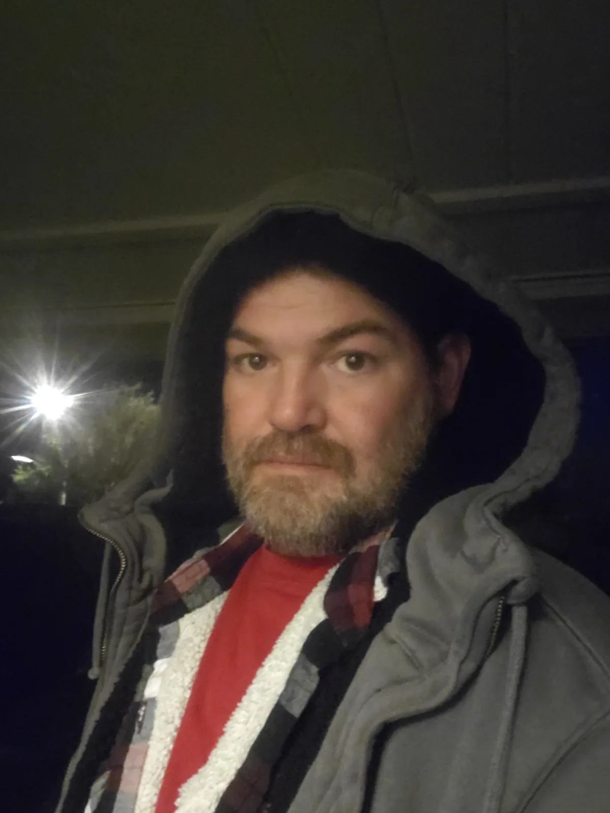 A man with a beard wearing a grey hooded jacket over a red shirt and plaid flannel, standing at night with a bright…