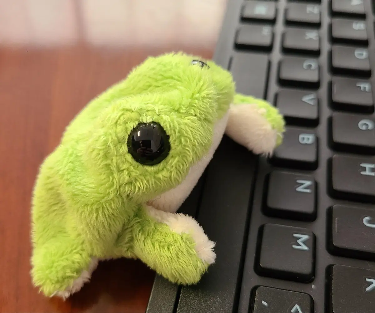 A bright green, fluffy plush frog with large black eyes is perched on the edge of a black computer keyboard, appearing to be…