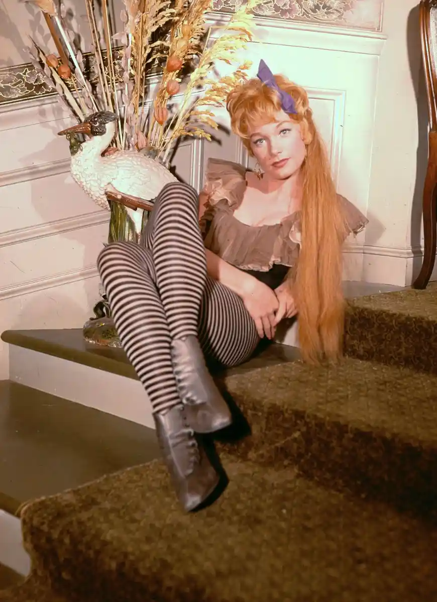 a woman with long red hair and a purple bow sits on carpeted stairs next to a decorative swan vase filled with dried…