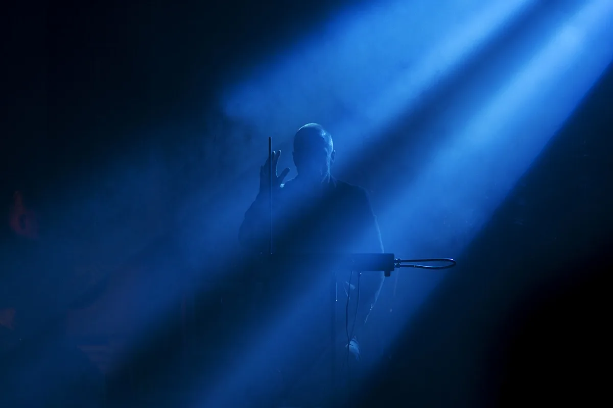 A bald man stands on a dimly lit stage bathed in blue light and smoke, holding a microphone stand with one hand while…