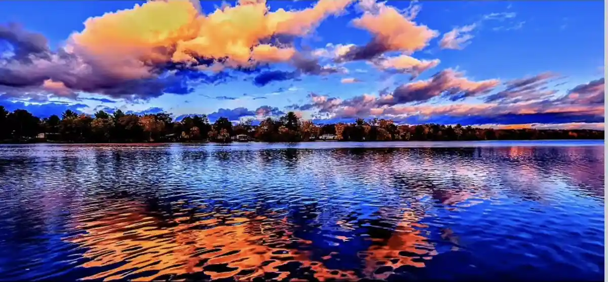 A vibrant, colorful sunset over a calm lake with trees on the horizon, where the sky's orange and blue hues reflect in the…