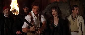 A still from a movie shows five characters standing in a dimly lit, rustic setting with a torch flame visible on the left…