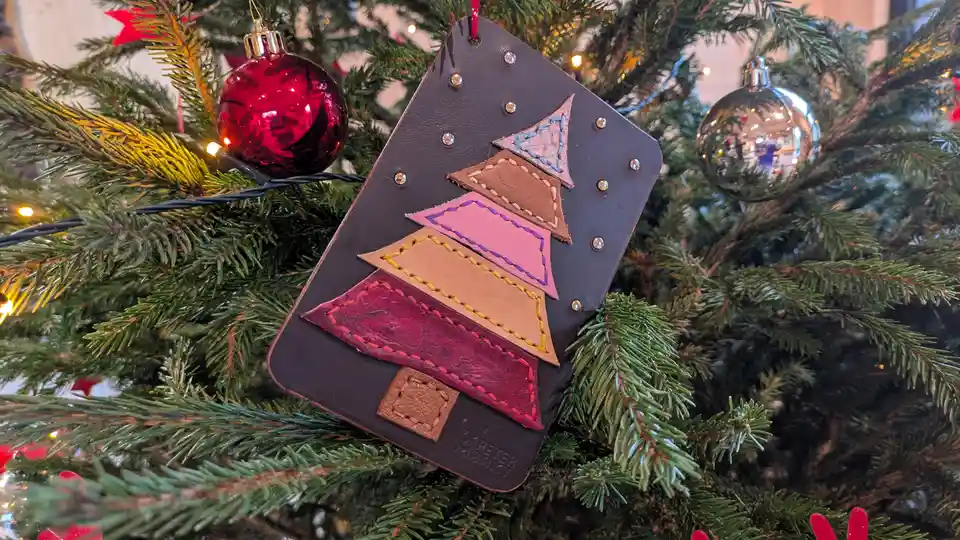 A handmade leather Christmas tree ornament, constructed from layered stitched leather pieces in pink, brown, and red, hangs…