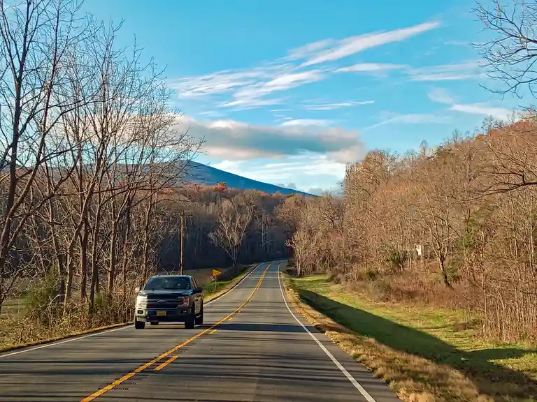 A black pickup truck drives down a winding rural road flanked by bare trees under a blue sky with wispy clouds. A distant…