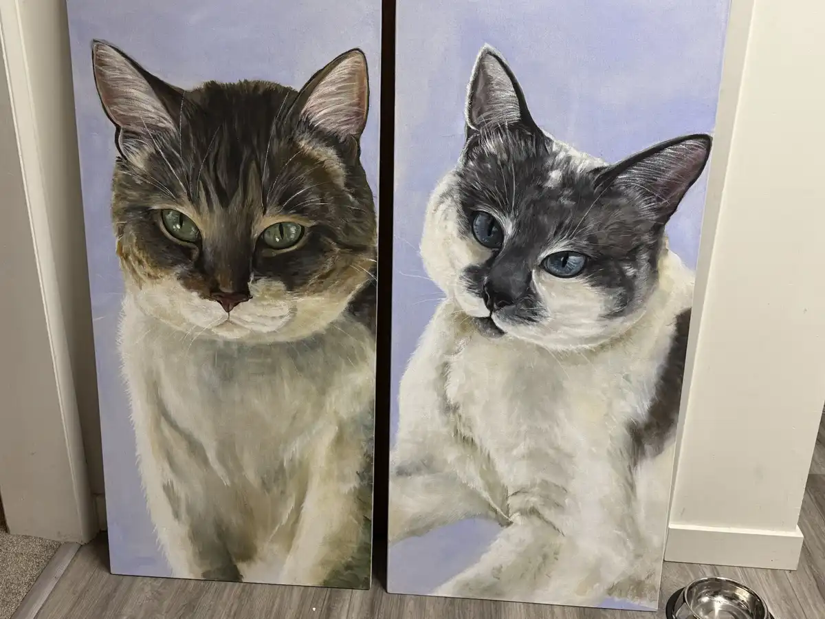 Two large, vertically oriented paintings of cats are standing side-by-side on a wooden floor, leaning against a wall. The…