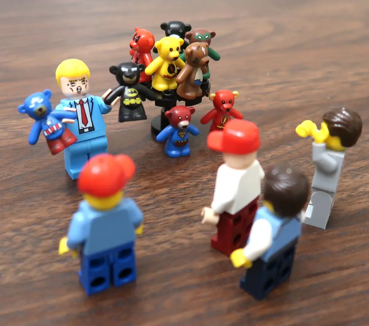 A Lego minifigure with blonde hair and a blue suit, holding a blue teddy bear, stands facing a group of other Lego figures…