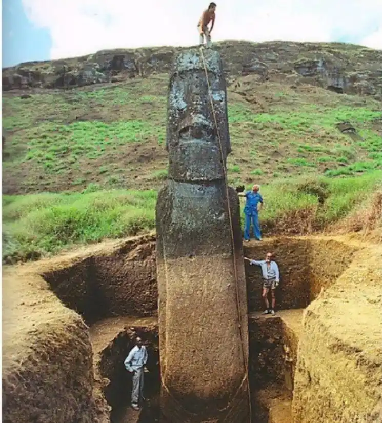A large, upright stone moai statue is partially unearthed from a deep excavation pit, with four men around it. One man…