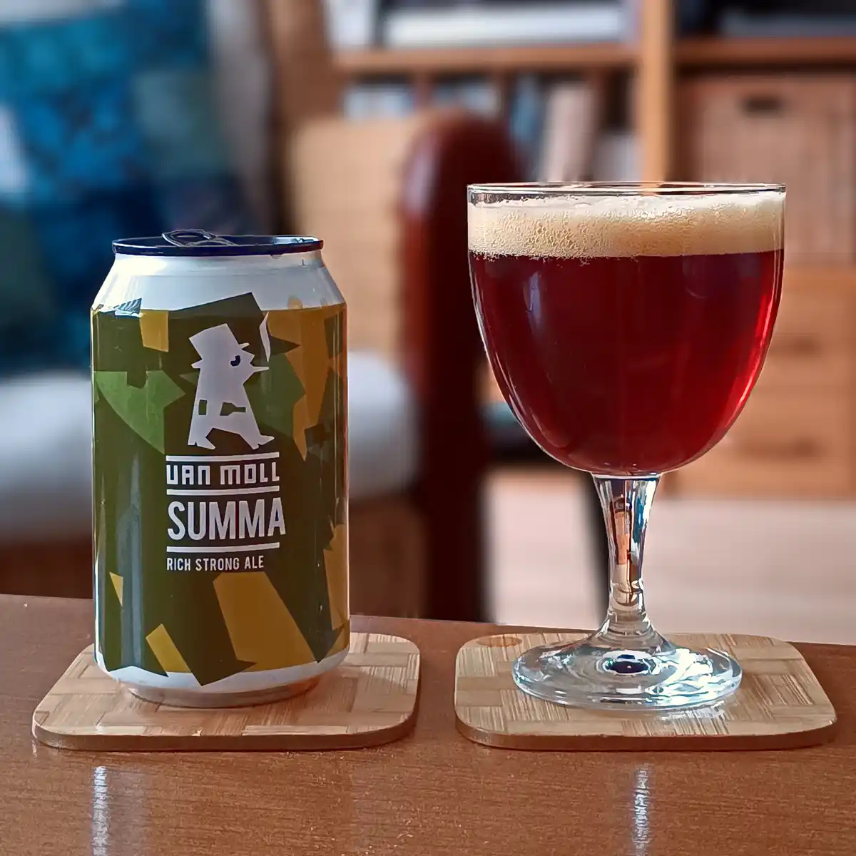 A can of Van Moll Summa Rich Strong Ale sits next to a stemmed glass filled with the dark red ale and topped with a light…